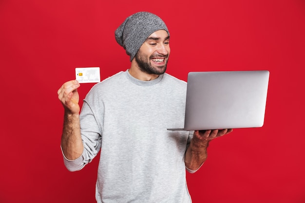 Foto van ongeschoren kerel 30s in vrijetijdskleding met creditcard en zilveren laptop geïsoleerd