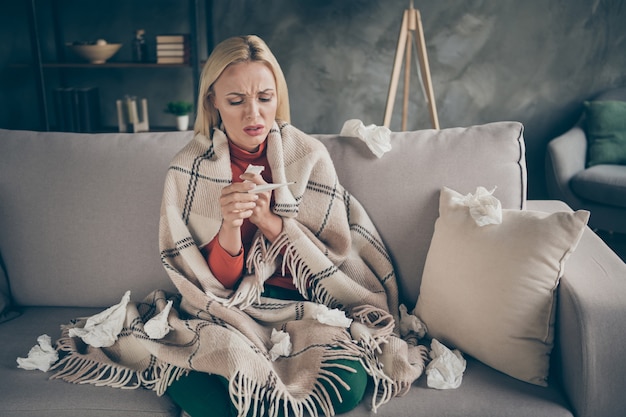 Foto van ongezonde mooie blonde dame die lijdt aan griep, gevangen koud papieren servetten overal houden thermometer zittend bank bedekt geruite deken woonkamer binnenshuis