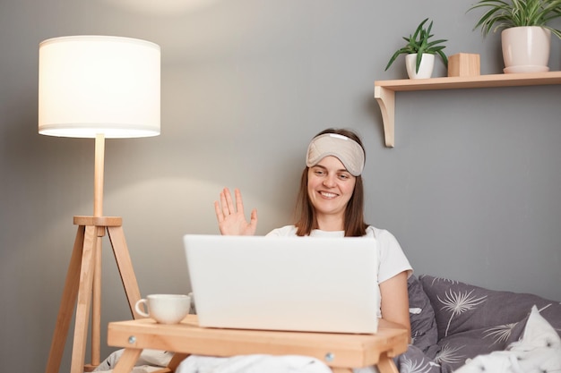 Foto van opgetogen charmante tevreden vriendelijke vrouw in wit t-shirt en blinddoek zittend in bed voor laptop en zwaaiende hand die hallo zegt met videogesprek