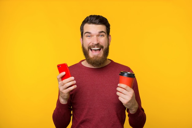 Foto van opgewonden bebaarde man in rode trui, glimlachend en camera kijken terwijl hij zijn mobiele telefoon en een kopje wit koffie