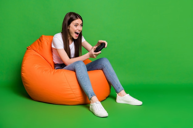 Foto van opgewonden grappige brunette haar jonge vrouw playstation slijtage t-shirt jeans sneakers zitten op poef geïsoleerd op groene achtergrond