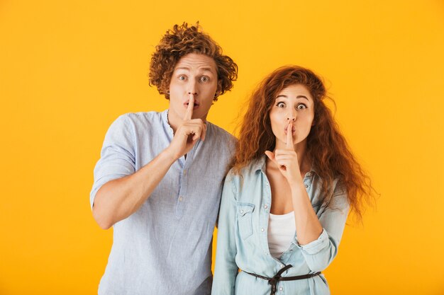 Foto van optimistische mensen, man en vrouw in basiskleding die en vinger bij mond glimlachen houden, die over gele achtergrond wordt geïsoleerd