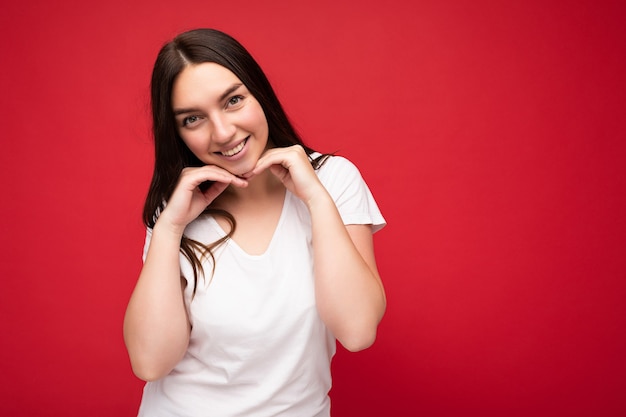 Foto van positief lachende jonge schattige mooie brunette vrouwelijke persoon met oprechte emoties, gekleed in casual wit t-shirt voor mockup geïsoleerd op rode achtergrond met kopie ruimte.