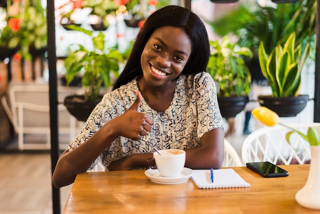 Foto van positieve donkere huid gemengd ras vrouw geniet van een goede nachtrust in de coffeeshop, drinkt warme dranken, heeft een brede glimlach, graag iets grappigs bespreken met vrienden. Mensen, vrije tijd en eten concept