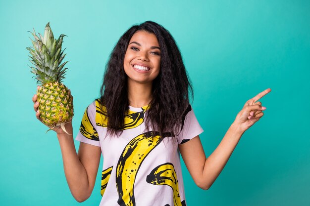 Foto van positieve donkere huid meisje punt vinger copyspace houd ananas draag roze t-shirt geïsoleerd op groenblauw kleur achtergrond