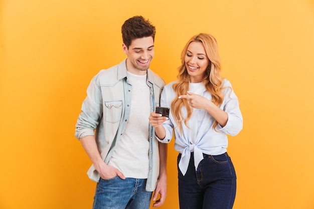 Foto van positieve mensen man en vrouw lachend terwijl gebaren op mobiele telefoon, geïsoleerd over gele muur