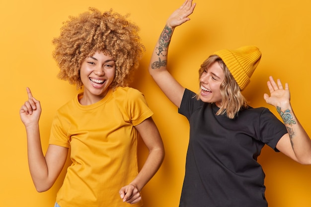 Foto van positieve twee jonge vrouwen schudden armen dans zorgeloos gekleed in zwarte en gele t-shirts voel me erg blij geïsoleerd over gele achtergrond chill samen veel plezier Gelukkig emoties concept