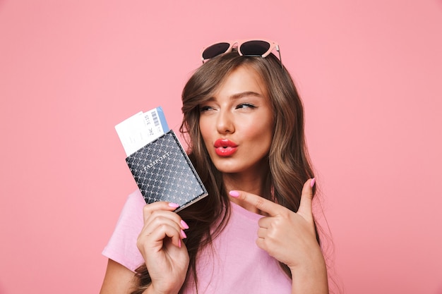 Foto van prachtige glamour vrouw met mooi lang haar wijzende vinger op paspoort en vliegtickets in de hand houden, geïsoleerd op roze achtergrond