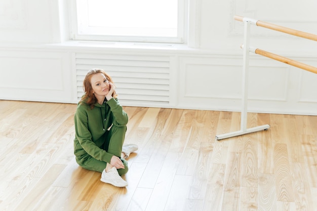 Foto van professionele danseres heeft rust na training poses op de vloer in de buurt van ballet barre heeft balletlessen draagt groene sweatsuit sportschoenen houdt ervan actief te zijn Mensen fitness en oefenen