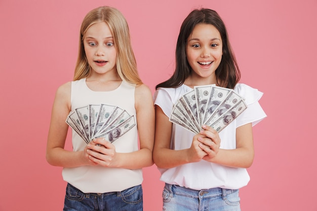 Foto van rijke brunette en blonde meisjes 8-10 die vrijetijdskleding dragen die veel geld in dollarbankbiljetten houden, die over roze achtergrond worden geïsoleerd