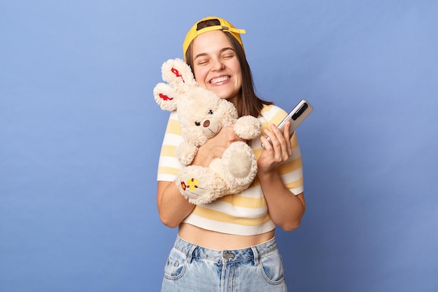 Foto van schattig charmant glimlachend tienermeisje met baseballpet en casual T-shirt staande geïsoleerd over blauwe achtergrond pluizig konijn speelgoed omarmend en mobiele telefoon vasthoudend