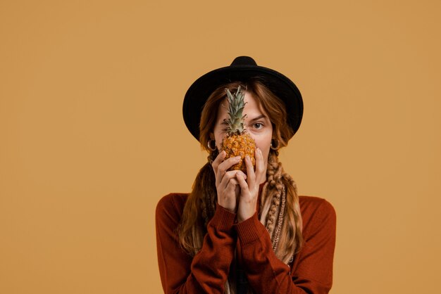 Foto van schattige boerin houdt ananas en bedekt met de voorkant naar boven. Blanke vrouw draagt denim overall en hoed geïsoleerde bruine kleur achtergrond.