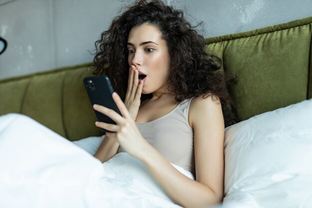 Foto van schattige geschokt mooie jonge vrouw binnenshuis thuis met behulp van mobiele telefoon chatten ligt in bed.