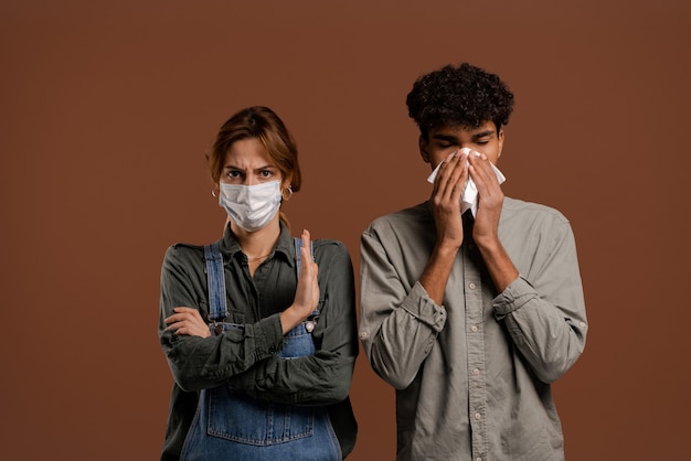 Foto van schattige paar boeren, vrouw met gezichtsmasker toont afstand terwijl man zijn neus snuit. Draagt denim overall, geïsoleerde bruine kleur achtergrond.