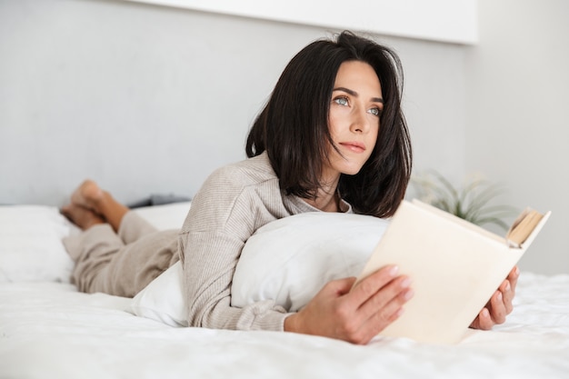 Foto van schattige vrouw 30s boek lezen, liggend in bed met wit linnen thuis