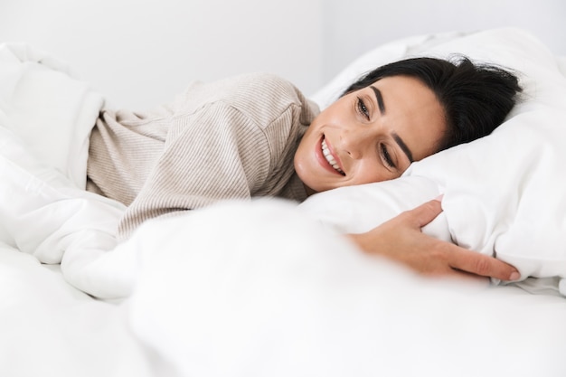 Foto van schattige vrouw 30s glimlachend, liggend in bed met wit linnen thuis