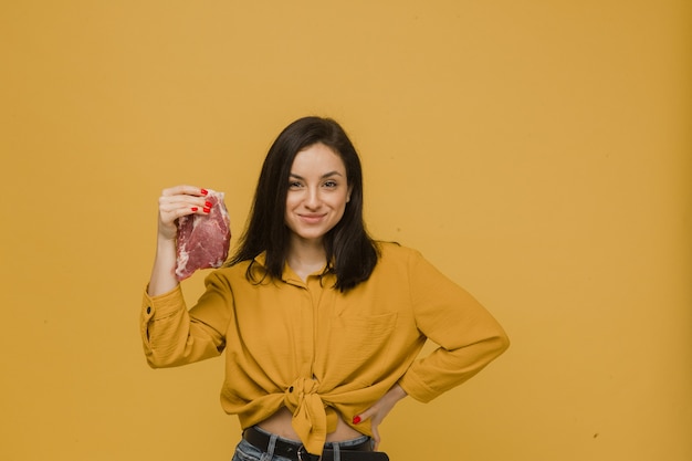 Foto van schattige vrouw houdt stuk vlees, vegetarische levensstijl. draagt geel shirt, geïsoleerde gele kleur achtergrond.