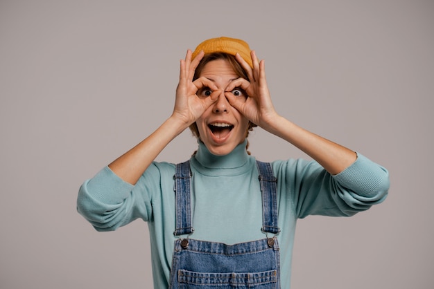 Foto van schattige vrouwelijke hipster pretendeert kijken in glazen. Blanke vrouw draagt denim overall en hoed geïsoleerde grijze kleur achtergrond.