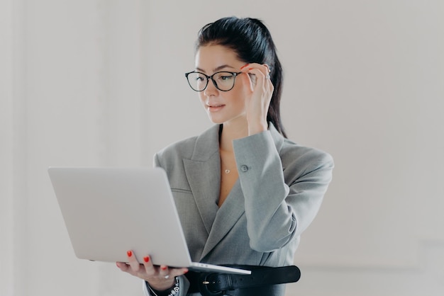 Foto van serieuze uitvoerende werknemer geconcentreerd in laptopscherm draagt transparante bril voor oogcorrectie bladert internet webpagina gekleed in elegante formele kleding staat tegen witte muur