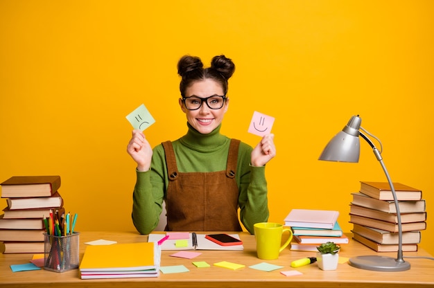 Foto van student dame houdt twee papieren kaarten emoticons zitten tafel op gele kleur achtergrond