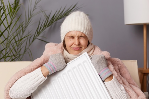 Foto van trieste depressieve vrouw in jas en hoed zit in een koude woonkamer, houdt de radiator in handen en worstelt thuis tegen koude bevriezing en huilt terwijl ze in een koud huis zit
