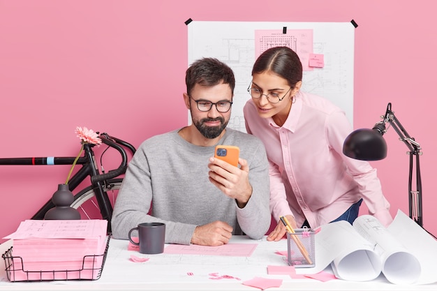 Foto van twee bekwame vrouw en man ontwerpers werken aan nieuw creatief project zie enkele voorbeelden van tekeningen in smartphone pose op werkplek spreek met elkaar geniet van samenwerking. Teamwerk concept