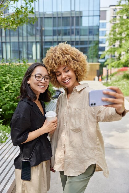Foto van twee gelukkige vrienden die afhaalkoffie drinken, selfie maken via mobiele pose tegen stedelijke achtergrond, gekleed in casual shirts, genieten van vrije tijd in de stad duizendjarige vrouwelijke reizigers nemen foto's