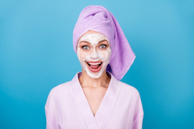Foto van verbaasde dame schuimmasker open mond glanzende glimlach draag violet handdoek tulband badjas geïsoleerde blauwe kleur achtergrond