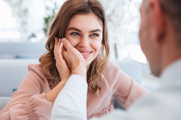 Foto van verliefde vrouw die op haar man kijkt en plezier neemt bij het houden van mannenhand op haar gezicht, terwijl ze een date heeft in een restaurant