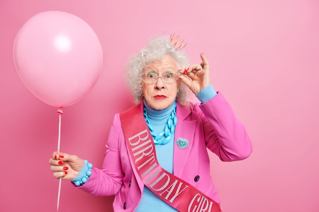 Foto van verwonderde gerimpelde vrouw houdt hand op de rand van een bril, gekleed in modieuze outfit houdt opgeblazen ballon viert verjaardag