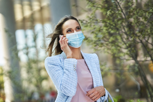 Foto van volwassen aantrekkelijke vrouw die een masker draagt met smartphone die in de stad loopt