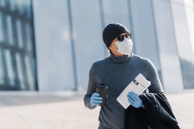 Foto van volwassen man loopt op straat, draagt beschermend masker en rubberen handschoenen als bescherming tegen besmettelijke ziekten, drinkt afhaalkoffie, kocht krant om nieuws te lezen, terzijde geconcentreerd