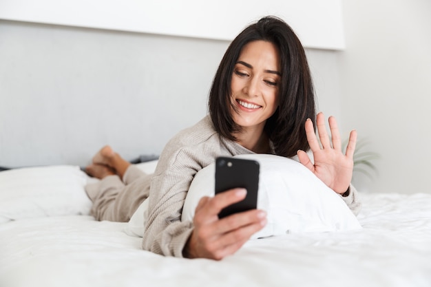 Foto van volwassen vrouw 30s met behulp van mobiele telefoon, liggend in bed met wit linnen in lichte kamer