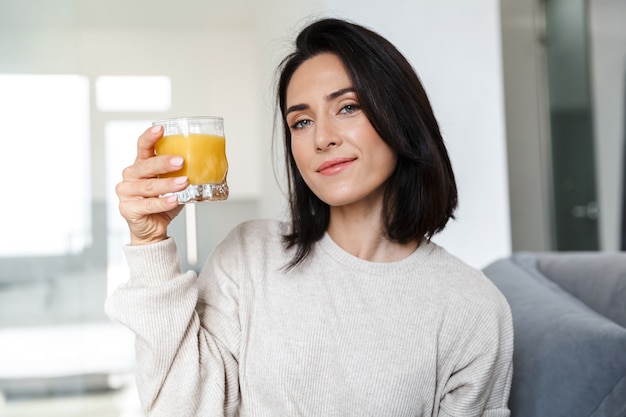 Foto van volwassen vrouw 30s sinaasappelsap drinken, zittend op de bank in lichte kamer