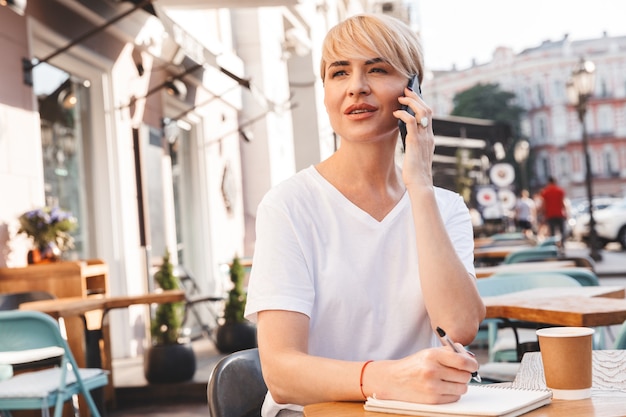 Foto van volwassen zakenvrouw dragen casual kleding zitten in zomerterras buiten, tijdens het opschrijven in notitieblok en spreken op mobiele telefoon