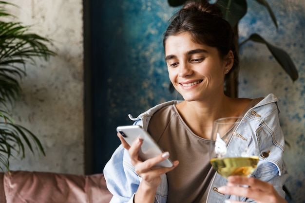 Foto van vrij mooie jonge vrouw zitten in café binnenshuis drinken van witte wijn met behulp van mobiele telefoon.