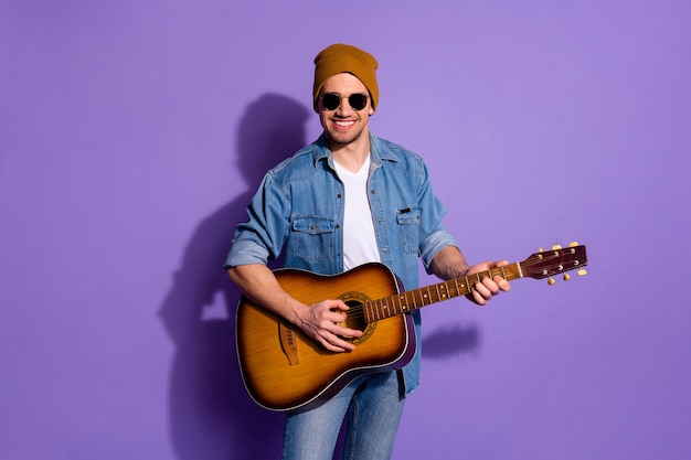 Foto van vrolijke aantrekkelijke trendy knappe coole aardige man met gitaar met hoofddeksels handen musicus instrument geïsoleerd op levendige paarse achtergrond te spelen