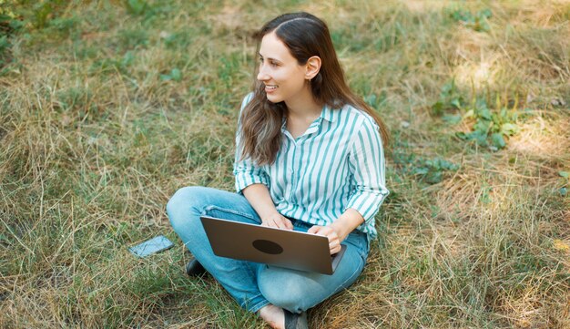 Foto van vrolijke jonge vrouw zittend op gras in park en met behulp van laptop.