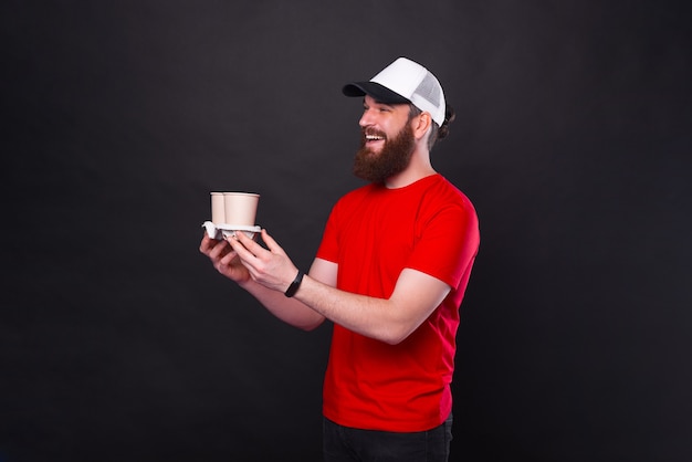 Foto van vrolijke man met baard die iemand twee koffiekopjes geeft om over zwarte achtergrond te gaan