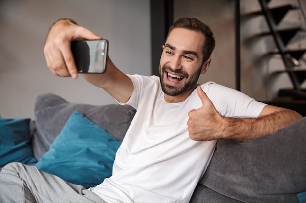 Foto van vrolijke vrijgezel 30s dragen casual t-shirt houden en nemen selfie op mobiele telefoon zittend op de Bank in de woonkamer