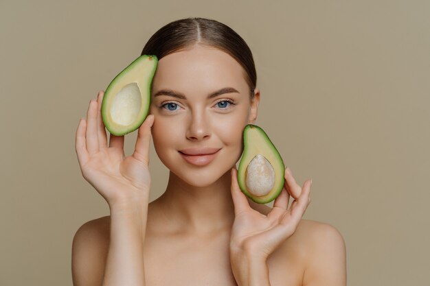 Foto van vrouwelijk model zonder shirt ziet er met zachte uitdrukking uit, houdt de helft van de avocado in de buurt van het gezicht heeft een gezonde, goed verzorgde huid na cosmetische behandelingen