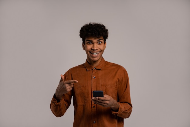 Foto van zwarte man houdt smartphones vast en wijst ernaar, ziet er erg verbaasd uit. Man draagt shirt, geïsoleerde grijze kleur achtergrond.
