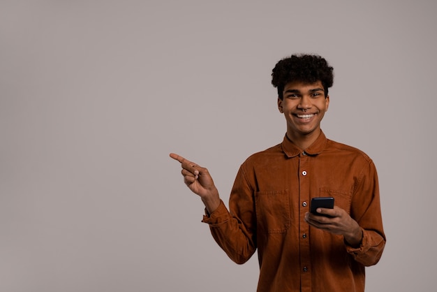 Foto van zwarte man houdt smartphones vast en wijst naar links voor advertentie, ziet er goed uit. Man draagt shirt, geïsoleerde grijze kleur achtergrond.