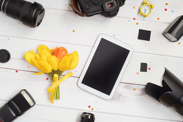 Fotograaf accessoires: camera, lens, flitser, geheugenkaart, wekker, tablet en een boeket tulpen op een houten tafel.