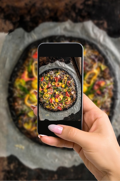 Fotograferen van voedselconcept - vrouw neemt foto van italiaanse pizza met zwart deeg en zeevruchten op een bakplaat uit de oven. Fotograferen op een mobiele telefoon of tablet