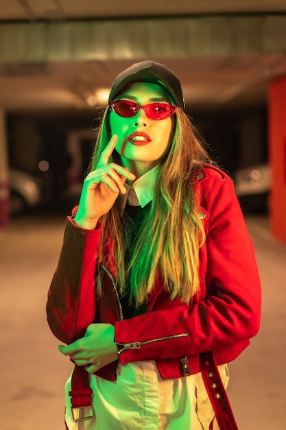 Fotografie met rode en groene neons op een parkeerplaats. Portret van een jonge vrij blonde blanke vrouw in een rood pak, zonnebril en een zwarte pet