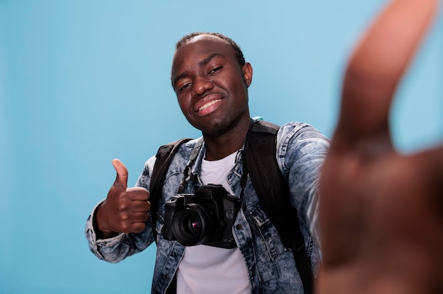 Fotografieliefhebber die een handgebaar geeft terwijl hij een foto van zichzelf maakt. Zelfverzekerde fotograaf die een goed vingersymbool toont terwijl hij selfie neemt en een DSLR-camera op een blauwe achtergrond heeft.