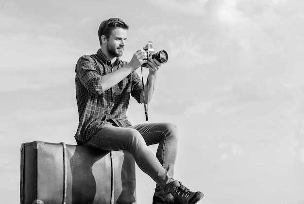 Fotojournalist concept Reizen met bagage Reisblogger Vlog schieten Vakantietijd Reisblog Man zit op koffer Knappe jongen reiziger retro camera Man buitenshuis met vintage koffer