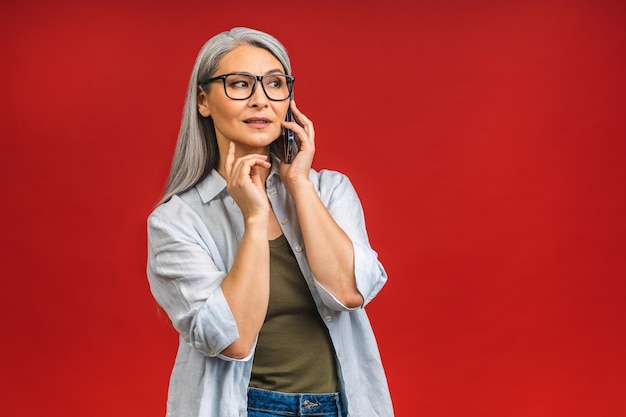 Fotoportret van gelukkige Aziatische senior volwassen oude vrouw die praat op de mobiele telefoon in casual geïsoleerd op rode achtergrond