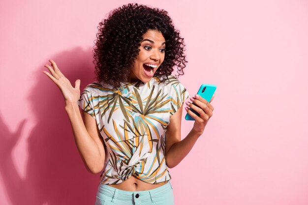 Fotoportret van geschokt schreeuwend meisje dat onverwacht een telefoon in de hand houdt, geïsoleerd op een pastelroze gekleurde achtergrond